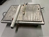 Full side view of the Rigid wet tile saw on the floor showing metal table with slots and saw blade.