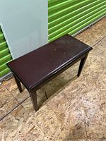 Angled view of dark brown wooden piano bench with cushioned top showing overall condition, surface wear visible on seat.