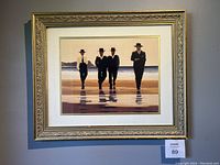 Full view of the framed art print showing four men walking on the beach, shadows reflected on wet sand, with gold ornate frame and cream matte border.