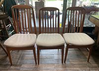 Three wooden dining chairs with vertical slats on the back and white padded cushions seen from the front against a shop window background.