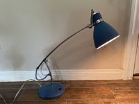 Blue adjustable desk lamp with curved arm and conical shade on wooden floor against white wall.