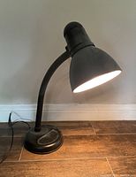 Black adjustable desk lamp standing on wood floor against a white wall. The lamp is switched on, showing the light inside the conical shade. The electrical cord is visible on the floor.
