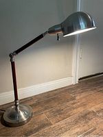 Full view of an adjustable desk lamp with metal rounded shade and wooden arm segments, turned on and casting light on floor.
