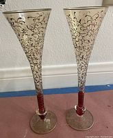 Two tall flared glasses with intricate gold vine swirl designs and red stems, placed upright on a floor against a white wall.
