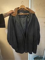 Photo of medium Cambridge brand jacket hanging on a wooden hanger showing black polyester material with brown collar and snap button zipper front closure.