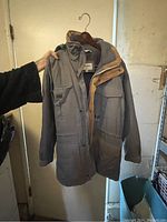 Front view of a Johns Bay men's jacket held on a hanger showing the full length, pocket details, and collar with brown suede accents