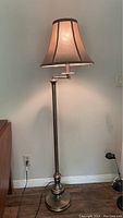 Photo of a metal floor lamp with a beige fabric shade turned on against a neutral wall.
