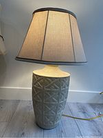 Photo of a ceramic table lamp with a beige fabric shade turned on, showing the geometric pattern on the base and lamp details.