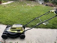 Side view of Ryobi electric lawn mower on concrete with grass in background, showing ergonomic handle and four wheels.