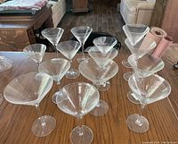 Photo showing 14 clear mismatched martini glasses arranged on a wooden table, illustrating variation in bowl shape and stem height.