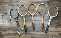 Top down photo of six vintage wooden tennis rackets on a wood floor, showing rackets in a row with varying sizes and grip colors.