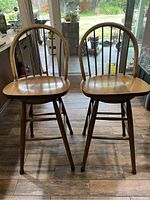 Photo of two wooden swivel bar stools with curved spindle backs and contoured wood seats on a wooden floor near a glass door with garden view.