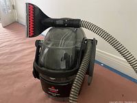 Photo of Bissell SpotClean Pro spot cleaner vacuum showing the main unit, clear dirty water tank, flexible hose, and brush tool attachment.