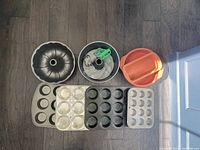 Photo showing various baking pans and molds on a wooden floor including bundt pans, round pans, loaf pans, and muffin tins of different sizes.