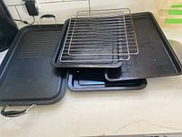 Photo showing a stack of three metal baking trays including a grill tray with handles and ridged surface, two flat baking sheets, and two metal cooling racks placed on top.