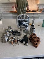Photo showing stainless steel cocktail shakers, boxed Rabbit wine tool set, silver textured napkin rings, wooden napkin holders, and assorted bottle stoppers and bar accessories on kitchen counter.