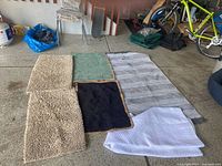 Four bath mats and one shower curtain displayed on garage floor. Mats include two beige shaggy, one green, one black. Shower curtain is white with horizontal silver-gray stripes.