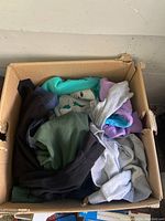 Top-down view of a box filled with folded adult hoodies in various colors including black, grey, purple, green, teal and blue.