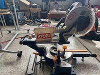 Side view of Ryobi compound miter saw showing blade, handle, dust bag, and miter adjustment controls.