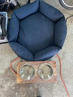 Photo showing a black circular dog bed and two stainless steel feeding bowls on a wooden stand placed on a concrete floor, with part of a bicycle visible.