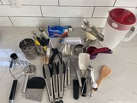 Photo showing assorted kitchen tools and gadgets arranged on a white tiled backsplash counter including knives, measuring cups, spoons, tongs, a rotary cheese grater, and a large plastic pitcher with red lid.