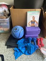 Exercise equipment including yoga blocks, dumbbells, weighted ball, stretch band, and workout book displayed on floor.