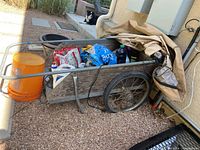 Photo of vintage wooden wheelbarrow filled with bags of fertilizer, weed killer, and orange gardening bucket, covered partially with a tarp.