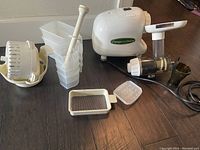 Photo shows white Omega juicer main motor unit with attached juicing section, electric cord, white pulp catcher, two translucent juice containers, plunger, fine mesh filter, and additional juicing parts on a wooden floor.