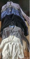 Seven men's dress shirts laid out flat, showing variety of colors and patterns including black, beige, gray, and plaid designs.