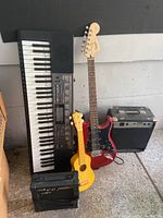 Full view of the lot showing electronic keyboard, yellow ukulele, red Fender-style electric guitar, and two guitar amplifiers.