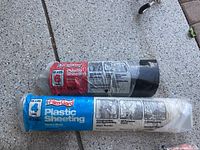 Two rolls of plastic sheeting on outdoor pavement. One roll is blue and white labeled Film-Gard Heavy Duty Plastic Sheeting 4 mil 5x25 ft, second roll red and black labeled Tough Plastic Sheeting 6 mil 10x25 ft.