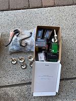 Box and bags with assorted hooks, hardware, screws, and nails, including black metal wall hooks, small brass hooks, and several boxes of small packaged hardware.