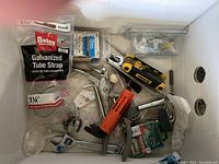 Top down view of multiple hand tools and hardware items including wrenches, pliers, utility knife and galvanized tube strap in plastic bag inside a box.
