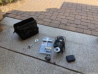 Shows Rotozip rotary tool with attached cutting bit, black soft carrying case, small wrench, packaged straight edge guide, extra circular cutting blade, and small black plastic box on concrete surface.