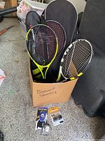 Sports gear including three tennis rackets in a box, two racket cases, two footballs on top, tennis grip and can of tennis balls on ground in front.