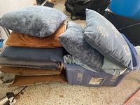 Stack of 9 assorted throw pillows in brown, gray, and blue colors next to plastic bin with more pillows inside, featuring textured fabrics and some patterned cases.