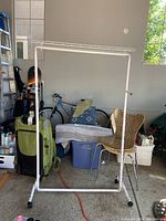 Two white metal hanging clothing racks shown against a garage background, with various household items around them.