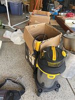 Stanley wet vacuum with stainless steel tank and yellow handle next to a used cardboard box containing various accessories including hoses and attachments