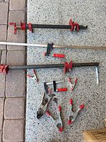 Photo showing a variety of woodworking clamps laid out on a speckled concrete floor and pavers. Includes two large red and black bar clamps, two smaller clamps with red handles, and multiple metal spring clamps with red rubber tips.