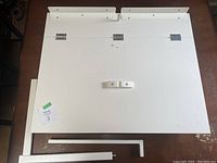 Overhead view of disassembled white drop-leaf table components including table surface and metal support pieces.