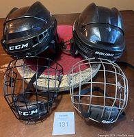 Both hockey helmets displayed from front angle, showing black CCM FM06 helmet on left and black BAUER True Vision II on right, with face cages detached and placed below the helmets.
