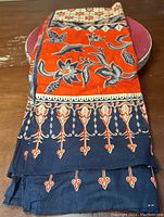 Folded Indonesian Batik tablecloth showing intricate orange and navy blue batik patterns, placed on a wooden table with a red round placemat beneath.