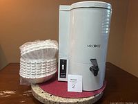 White Mr. Coffee 24 cup drip coffee maker with black spigot, water reservoir, and red water level indicator. Coffee filters wrapped in plastic sit next to the coffee maker on a red placemat.