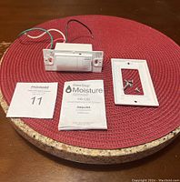 DewStop Moisture Sensing Bathroom Fan Control (Model HS-C00) displayed with wall plate, screws, and user manual on a red placemat with cork trivet beneath.