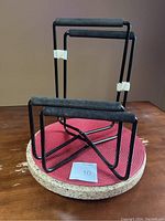 Two foldable black metal low profile speaker stands shown standing upright on a round surface with a reddish cloth mat underneath.