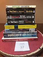 Collection of nine spy and espionage-themed books stacked on a red circular mat, showing spines and covers with titles and authors visible.