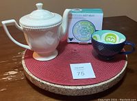 Ivory white Bowring Ivory Pearl porcelain teapot next to Native Home porcelain art mug with its box on round red and cork placemat on wooden table.