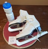 Photo of woman’s white figure skates size 8 with white blade guards placed on a red round mat next to a white water bottle with a blue top.