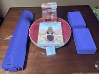 Set displayed on table including purple rolled yoga mat, two purple yoga blocks, Complete Yoga & Pilates workout DVD, Namastay yoga book, and a red round mat underneath the book and DVD.