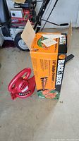 Photo showing a red electric leaf blower placed beside a Black and Decker 14 inch hedge trimmer boxed in orange and black packaging on a concrete or cement floor.